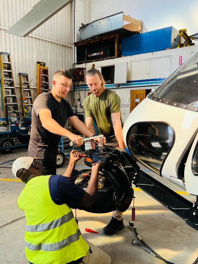 Ground crew fitting a stabilised camera gimbal to the nose of a helicopter in a hangar before an aerial filming charter