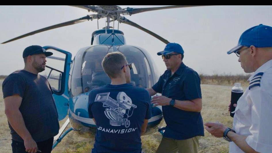 Aviation ONE pilot briefing a production camera crew beside a helicopter on a dirt airstrip before an aerial sortie