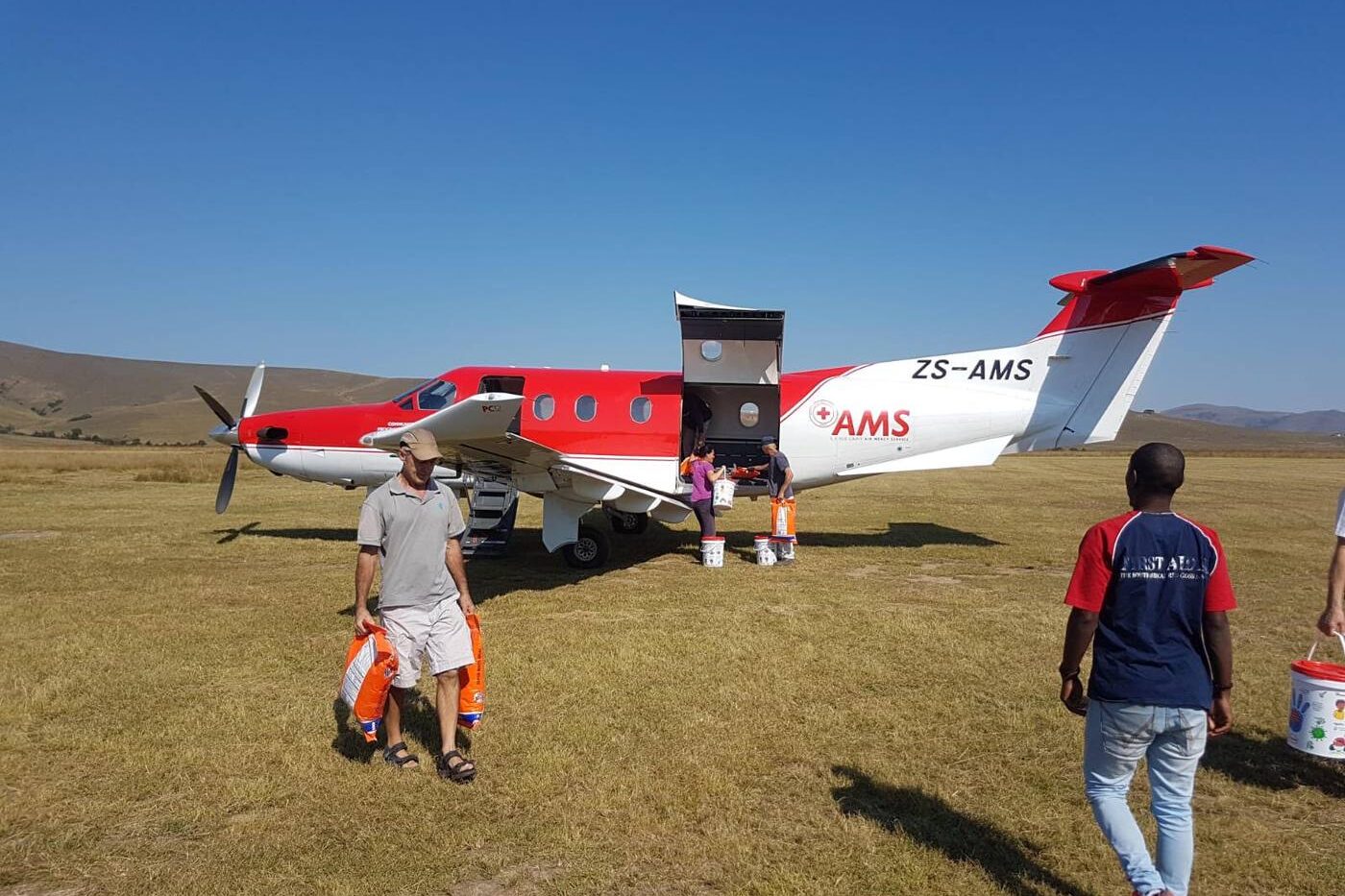 Pilatus PC-12 in air mercy service livery unloading relief supplies by hand on a rural grass airstrip